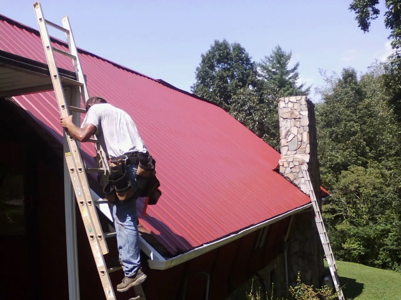 Roofer on a ladder beside a red metal roof and stone chimney — Soffit & Fascia Repair by Roofing Brothers in Nortonville